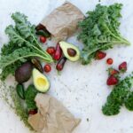Top view of fresh avocados, kale, tomatoes, and peppers arranged on a marble surface.