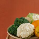 Close-up of fresh broccoli, cauliflower, lemon, and orange on a wooden plate with an earthy background.