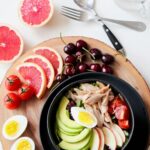 A vibrant breakfast flat lay featuring a mix of fruits, avocado, eggs, tomatoes, and sliced meats on a wooden platter.
